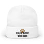 A white knit beanie hat with a folded cuff, featuring the text 'Life Is Better With Dogs' in black next to a brown paw print graphic.