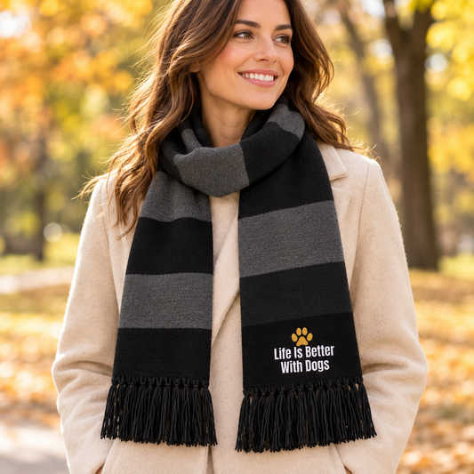 thick striped knit scarf in black and gray with fringe ends. The black section on the lower right of the scarf has white text reading 'Life is Better With dogs' and a small gold paw print logo