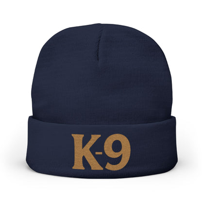 A navy blue knit beanie with a folded cuff featuring the text 'K-9' embroidered in a tan/gold color, shown against a plain white background.