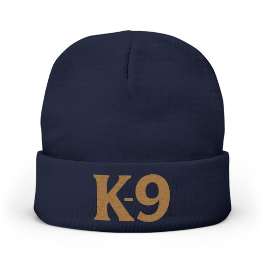 A navy blue knit beanie with a folded cuff featuring the text 'K-9' embroidered in a tan/gold color, shown against a plain white background.